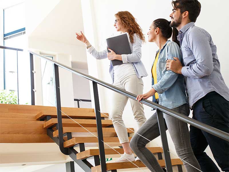 Frau steht mit jungem Paar auf einer Treppe und zeigt den potentiellen Mietern eine Immobilie.