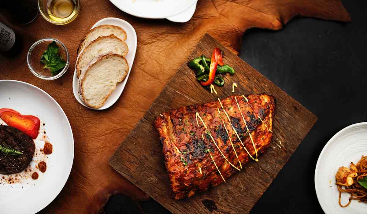 Eine frisch gegrilltes Stück Fleisch liegt auf einem Holzbrett mit Brot und Grillsoßen als Beilagen auf dem Tisch.