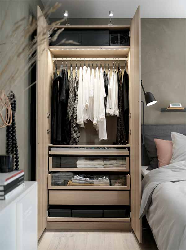 Ein geöffneter deckenhoher Kleiderschrank neben einem Bett, die Kleidung hängt und liegt sehr geordnet darin.